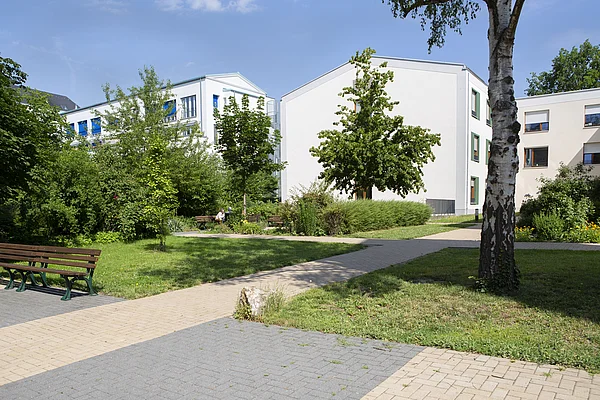 Außenanlage des Nellinistift  mit Sitzbänken auf barrierefreiem, gepflasterten Weg, umgeben von Rasen, Büschen und Bäumen, im Hintergrund Wohngebäude