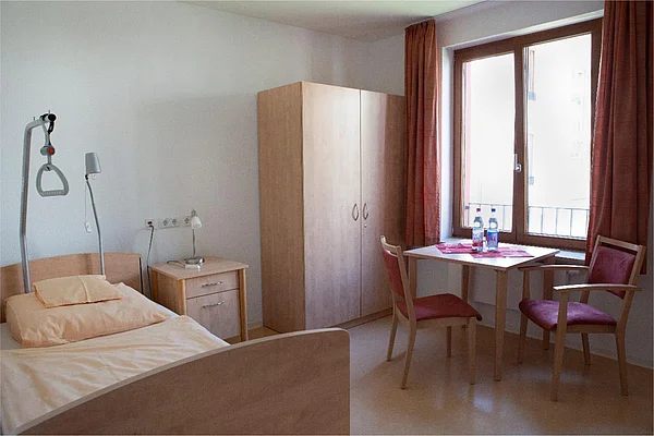 Zimmer mit Pflegebett, Nachttisch, Tisch, Kleiderschrank und Fenster