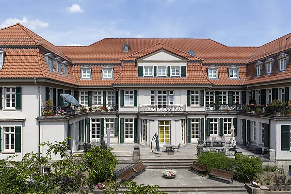 Großes, mehrstöckiges U-förmiges Gebäude mit Balkons, und einem Vorplatz mit Sitzgelegenheiten