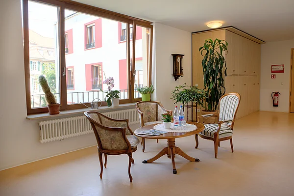 Runder Holztisch mit 3 Stühlen vor einem großen Fenster, im Hintergrund Pflanzen und ein großer Wandschrank